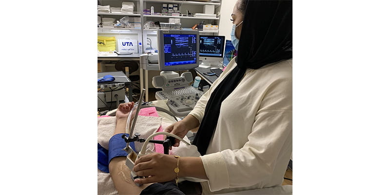 A healthcare professional, wearing a hijab and a white coat, is administering an ultrasound-guided procedure to a patient's arm. The patient's arm is extended, with a blue tourniquet applied, and the healthcare professional is holding an ultrasound probe and a syringe. In the background, an ultrasound machine with a live screen displaying data can be seen.